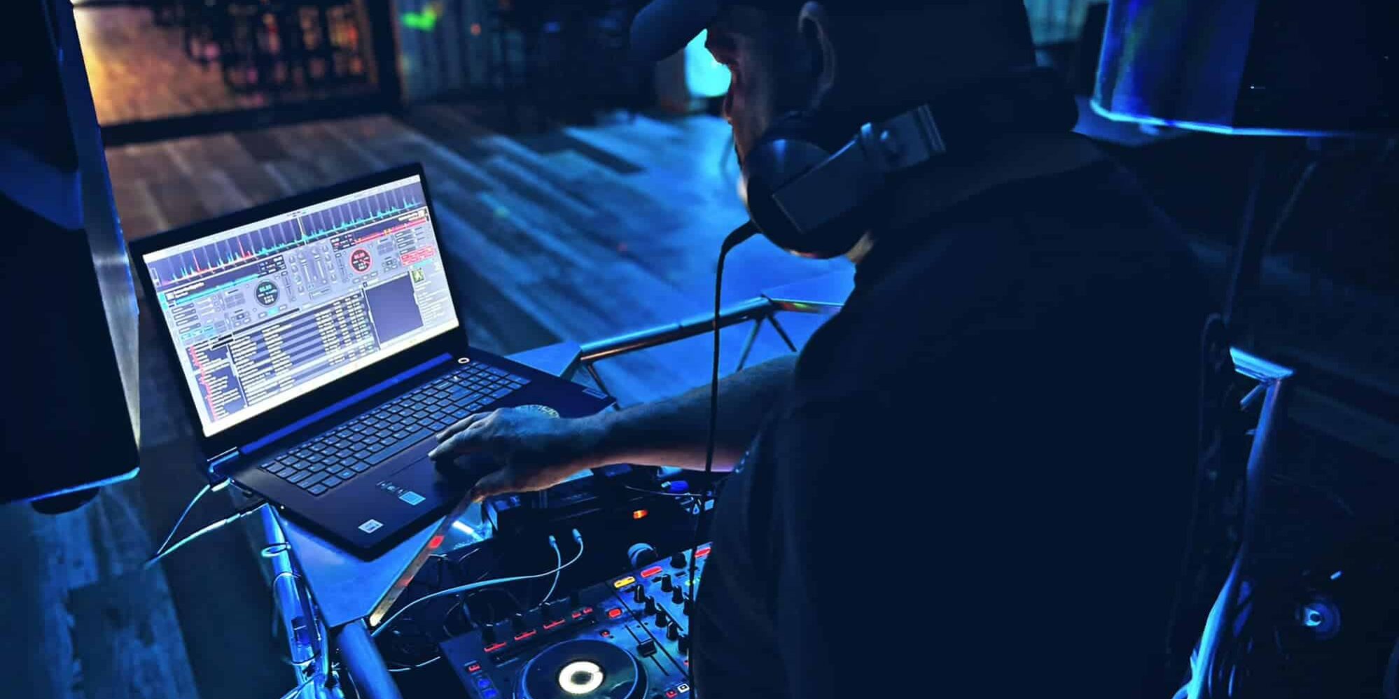 Photograph of a DJ performing at an event. The DJ, wearing a dark cap and headphones around their neck, is operating a laptop and a DJ controller with colorful lights in a dimly lit venue. The background shows a wooden floor and a few tables, with blue and purple lighting creating a lively atmosphere.