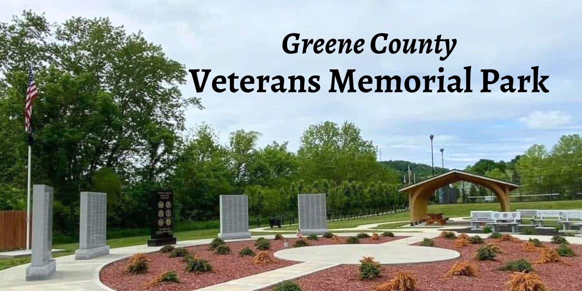 Greene County Veterans Memorial Park featuring a circular plaza with engraved memorial stones, an American flag, landscaped flower beds, and a covered wooden pavilion surrounded by trees.