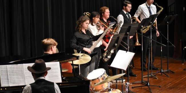 Student jazz ensemble performing on stage with piano, drums, bass, trombone, trumpet, and saxophones.
