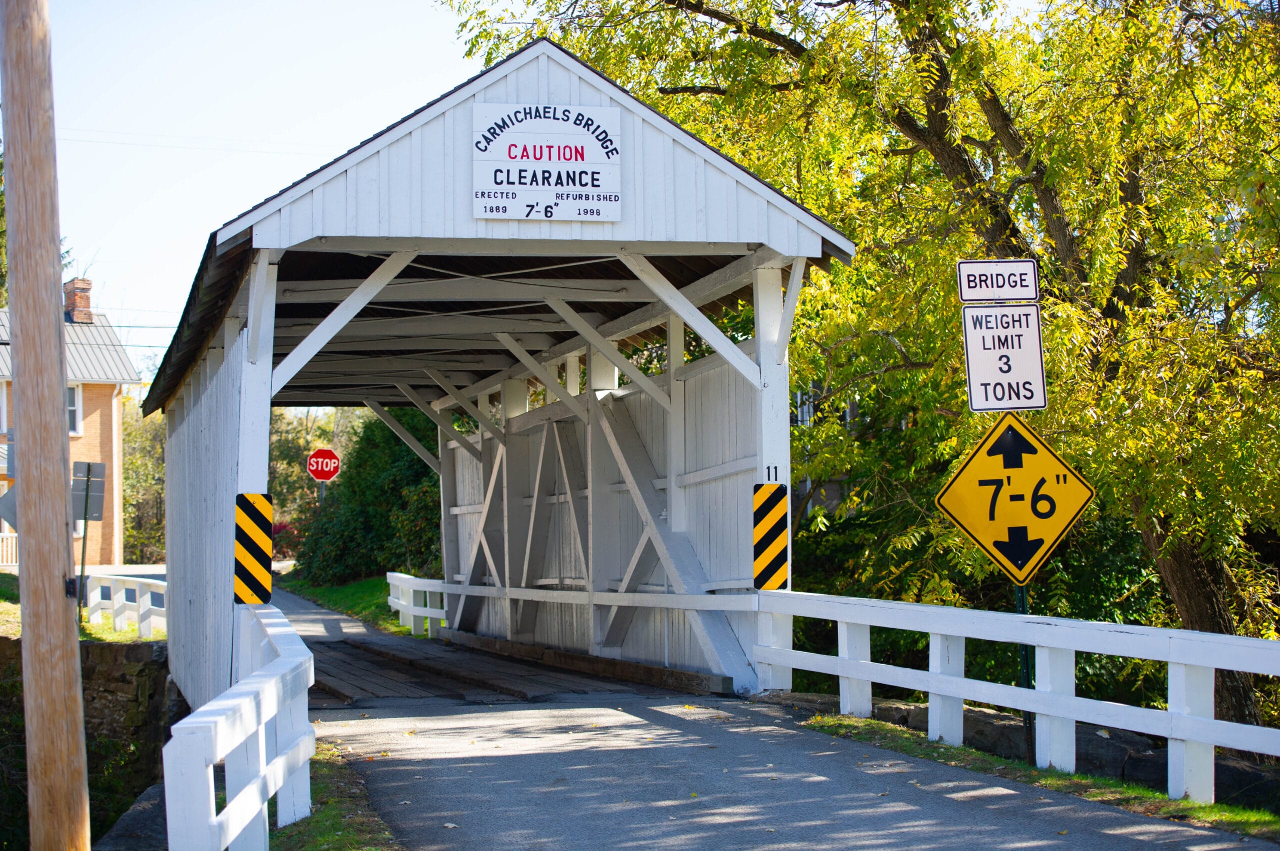 White covered bridge on a narrow road with clearance and weight limit signs, framed by green and yellow autumn trees on a sunny day