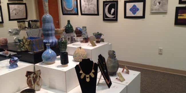 Photograph of student art exhibition at Waynesburg University with jewelry and pottery in the foreground. 2-D artwork is hanging on the wall.
