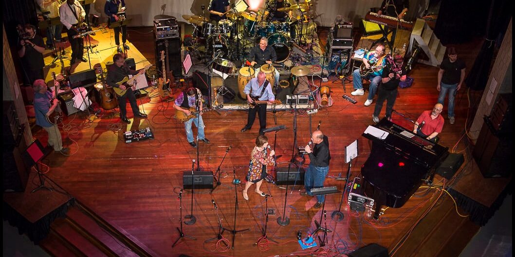 A large jazz band performing on a wooden stage. The ensemble includes multiple guitarists, saxophonists, a pianist, keyboardist, percussionists, and a drum set. Two vocalists sing at the front, surrounded by microphones and stage monitors. Colorful stage lighting highlights the musicians.