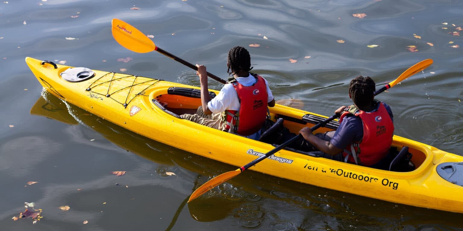 Two people wearing red life jackets paddling a yellow tandem kayak on calm water with scattered autumn leaves