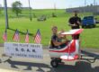 A woman and a young child ride in a small red-and-white airplane-shaped cart, smiling and waving, while a man stands behind them on a grassy field. A sign on the cart reads “The Plane Train by S.O.A.R. of Greene County,” decorated with several American flags.