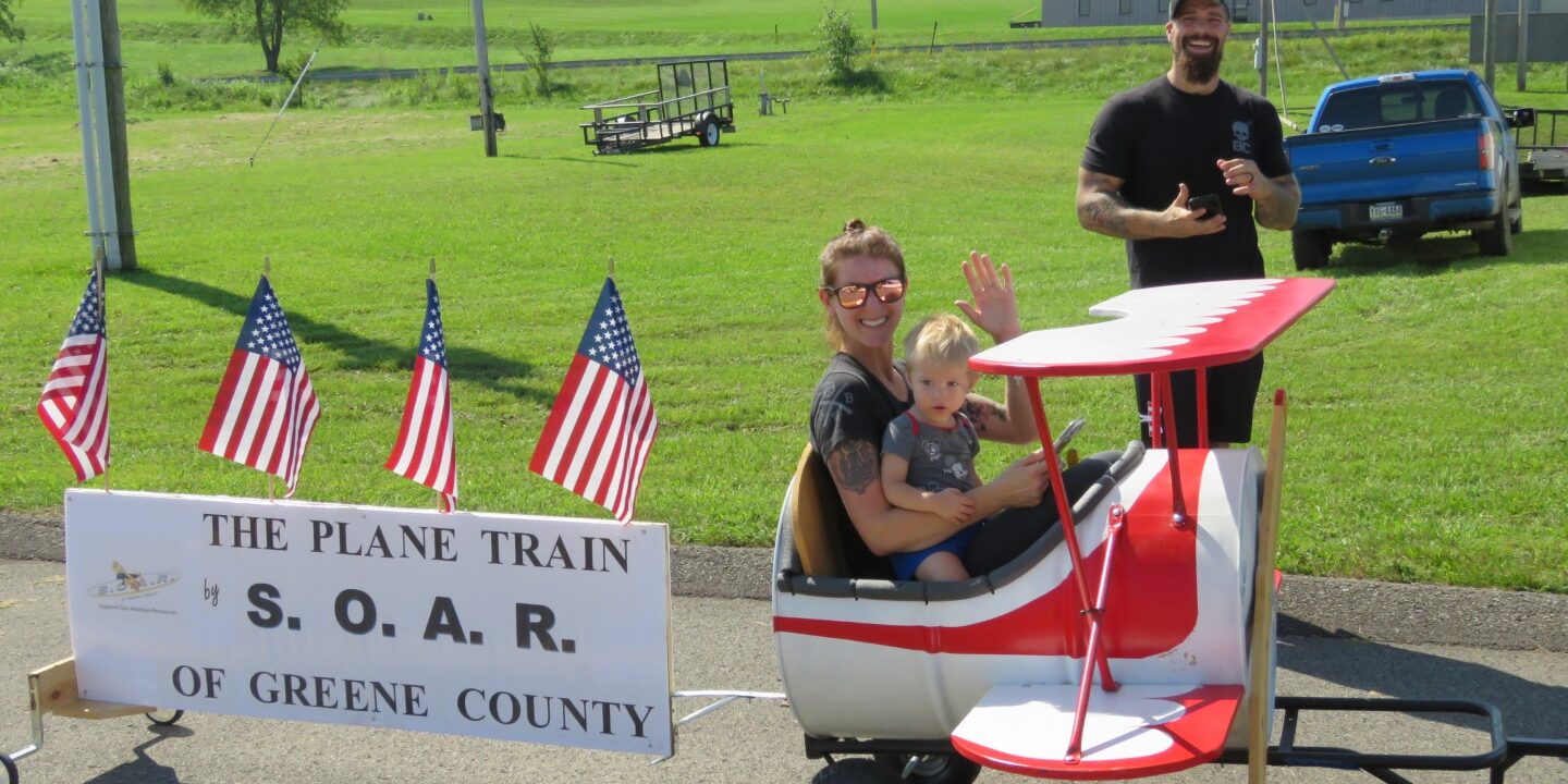 Aviation Day Plane Train A woman and a young child ride in a small red-and-white airplane-shaped cart, smiling and waving, while a man stands behind them on a grassy field. A sign on the cart reads “The Plane Train by S.O.A.R. of Greene County,” decorated with several American flags.