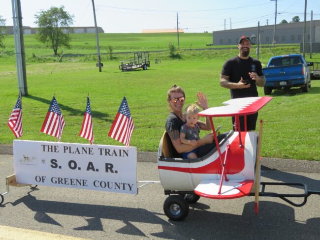Aviation Day Plane Train A woman and a young child ride in a small red-and-white airplane-shaped cart, smiling and waving, while a man stands behind them on a grassy field. A sign on the cart reads “The Plane Train by S.O.A.R. of Greene County,” decorated with several American flags.