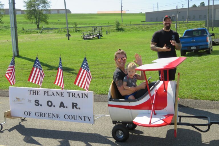 A woman and a young child ride in a small red-and-white airplane-shaped cart, smiling and waving, while a man stands behind them on a grassy field. A sign on the cart reads “The Plane Train by S.O.A.R. of Greene County,” decorated with several American flags.