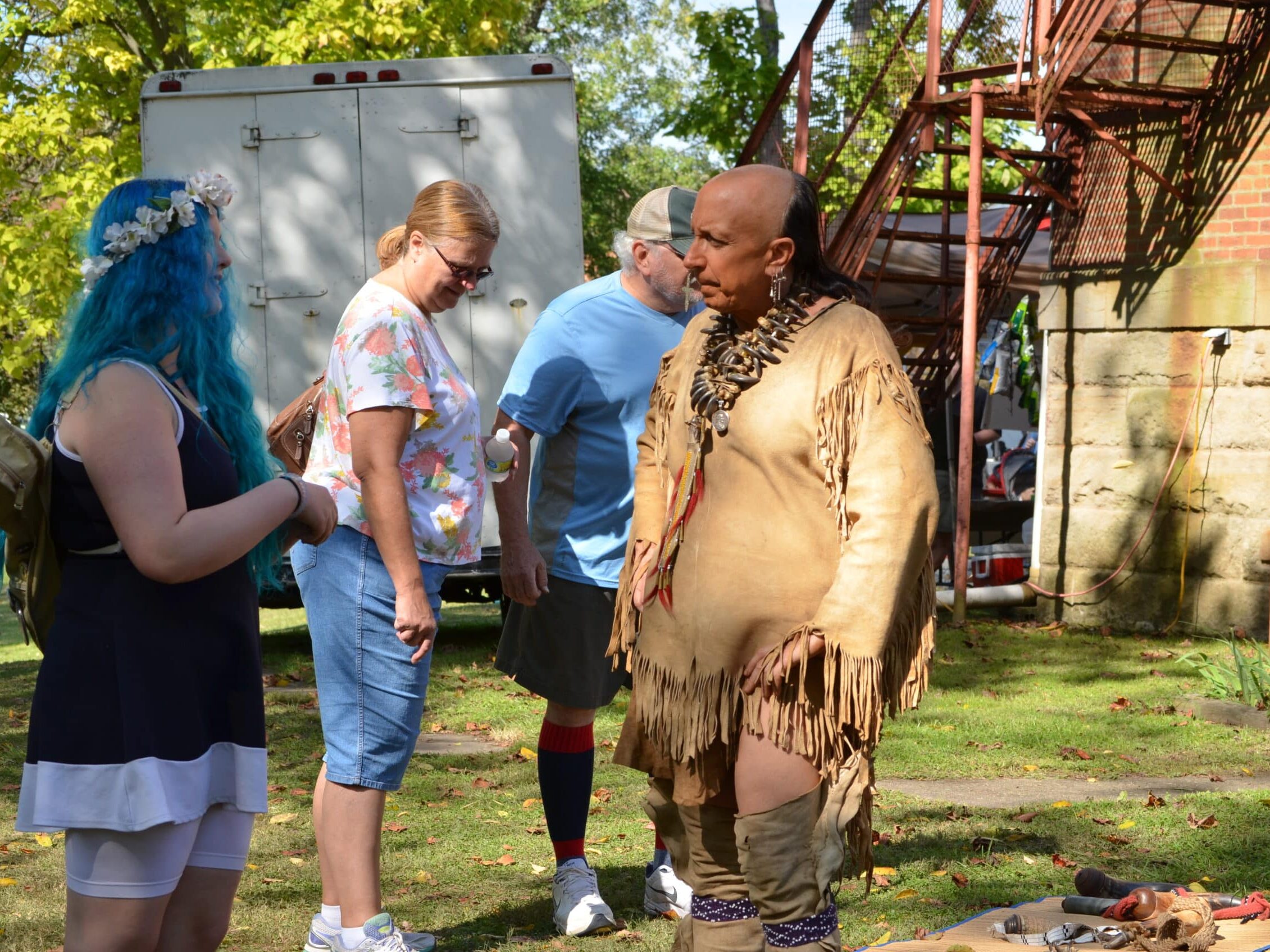 A small group of people stand outdoors on a grassy area beside a historic building, listening to a presenter in traditional buckskin clothing and necklace, with items displayed on a mat nearby.