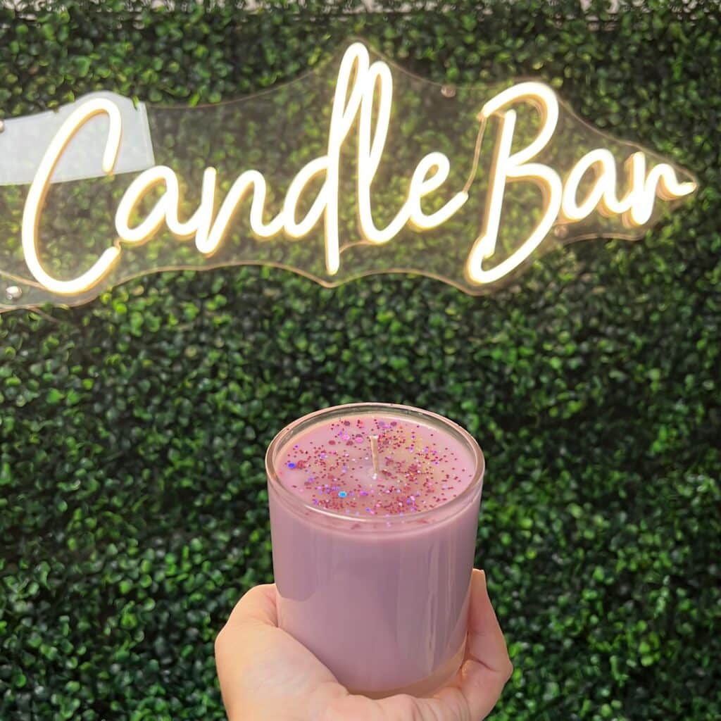 Photograph of a pink glitter candle in front of a wall of green ivy with lighted sign saying Candle Bar.
