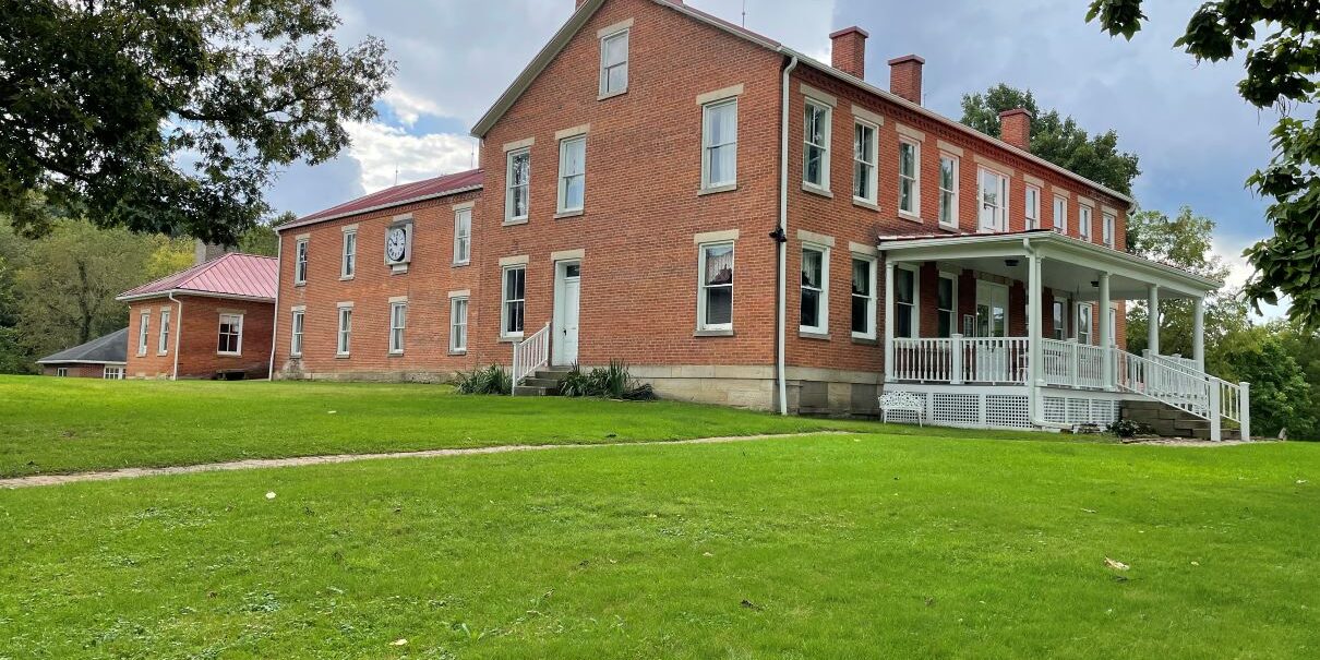 Large historic red-brick building with white trim and a covered front porch, set on a grassy lawn under a cloudy sky.
