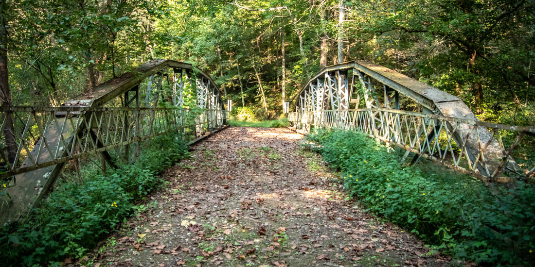 A narrow, weathered metal bridge with decorative railings spans a wooded area, surrounded by dense green trees and foliage. The dirt path across the bridge is scattered with fallen leaves, creating a quiet, rustic outdoor setting