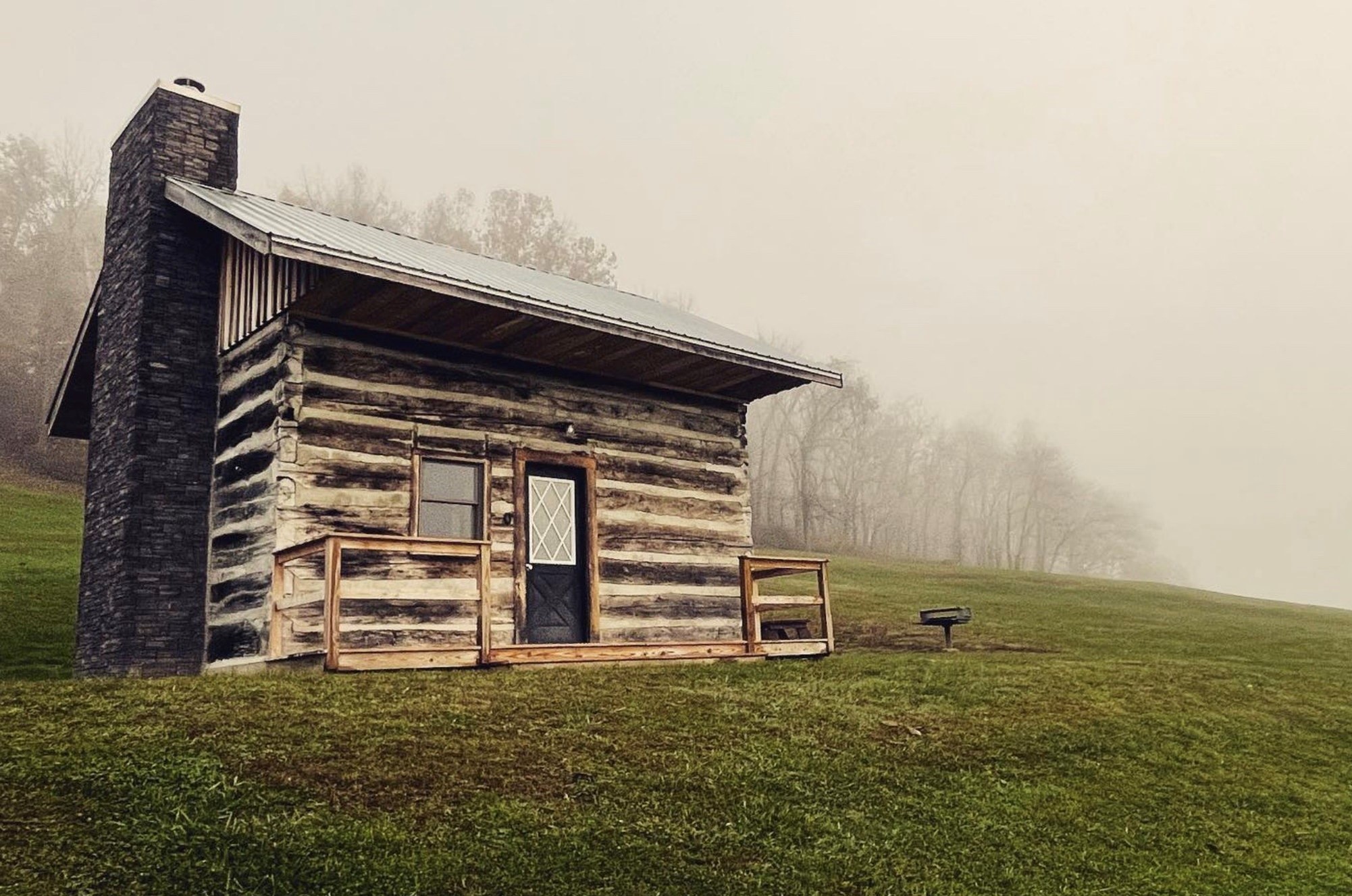 Small rustic log cabin with a stone chimney and metal roof, sitting on a grassy hillside in a foggy landscape with trees in the background