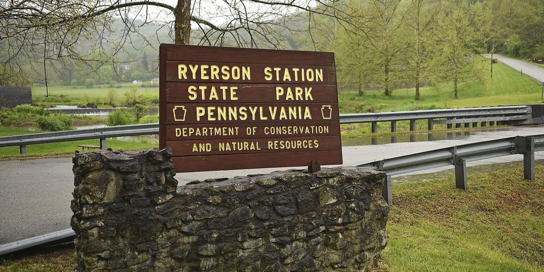 Wooden roadside sign for Ryerson Station State Park in Pennsylvania, mounted on a stone base, with a rural landscape of trees, grass, and a road in the background on a cloudy day.