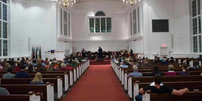 Audience seated in wooden pews inside a bright, white church sanctuary, facing a small orchestra performing at the front beneath chandeliers.