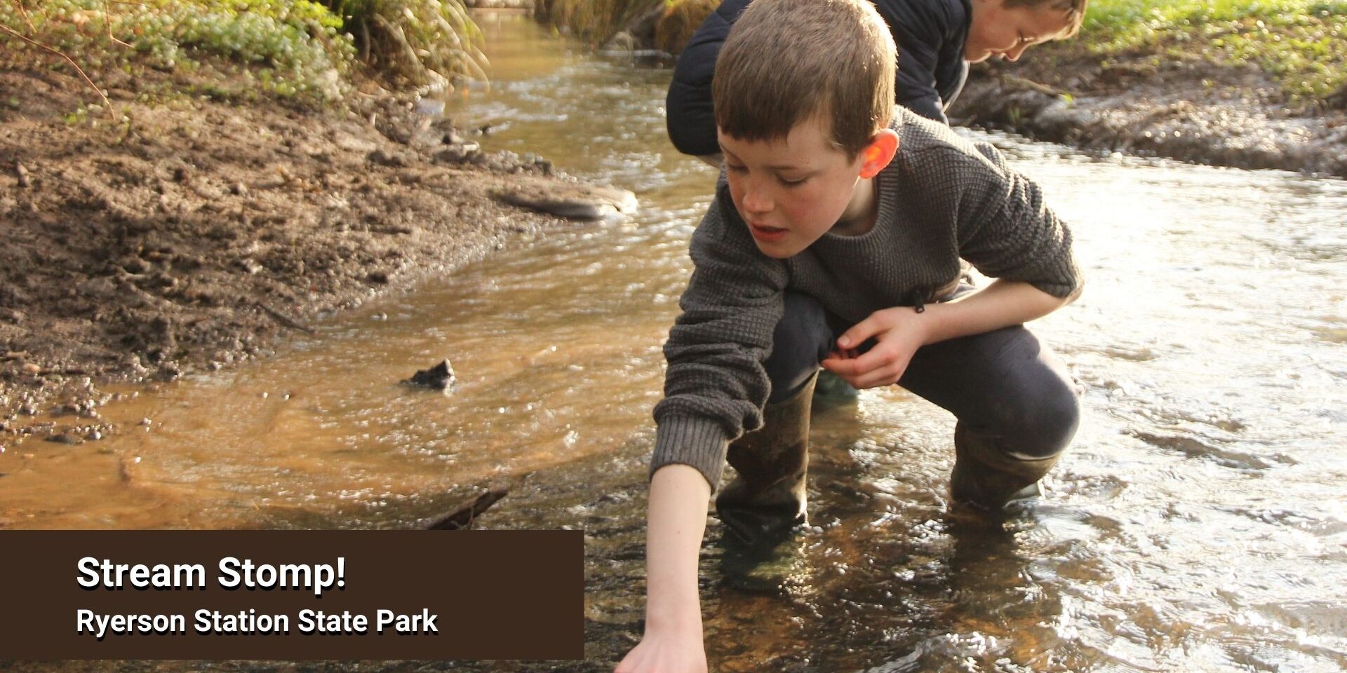 Two children wearing boots explore a shallow stream, crouching and reaching into the water, with text overlay reading “Stream Stomp! Ryerson Station State Park.”