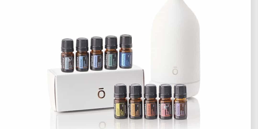 A white essential oil diffuser next to a boxed set of small amber glass bottles containing various essential oils, neatly arranged in two rows on a clean white background