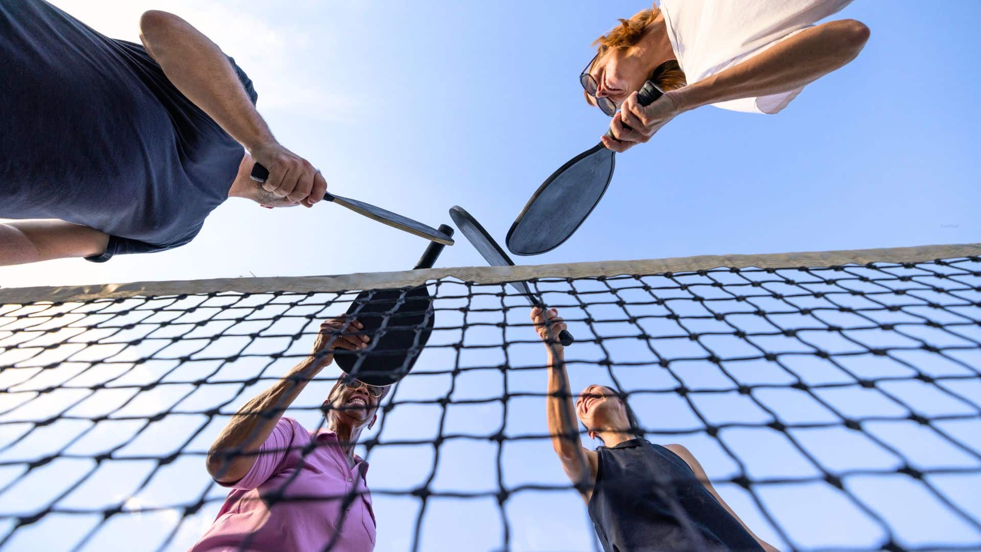 Photograph from the ground looking up at 4 pickleball players touching their racquets together.