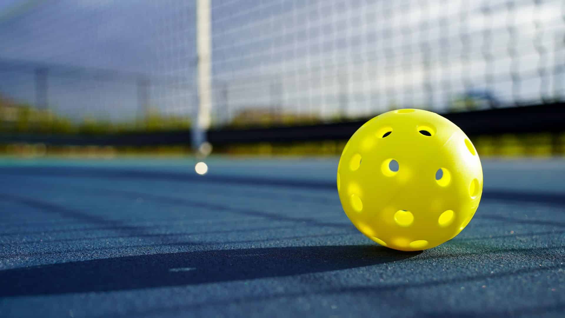 Photograph of a yellow pickleball laying on the blue court next to the net.