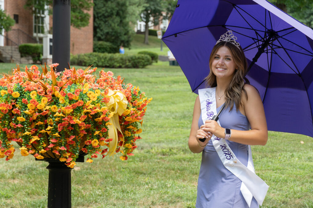 A young woman wearing a crown and a “Miss Rain Day 2025” sash stands outdoors holding a purple umbrella beside a large planter of orange and yellow flowers on a grassy lawn.