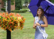 A young woman wearing a crown and a “Miss Rain Day 2025” sash stands outdoors holding a purple umbrella beside a large planter of orange and yellow flowers on a grassy lawn.