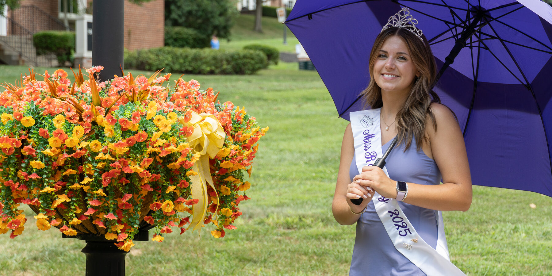 A young woman wearing a crown and a “Miss Rain Day 2025” sash stands outdoors holding a purple umbrella beside a large planter of orange and yellow flowers on a grassy lawn.