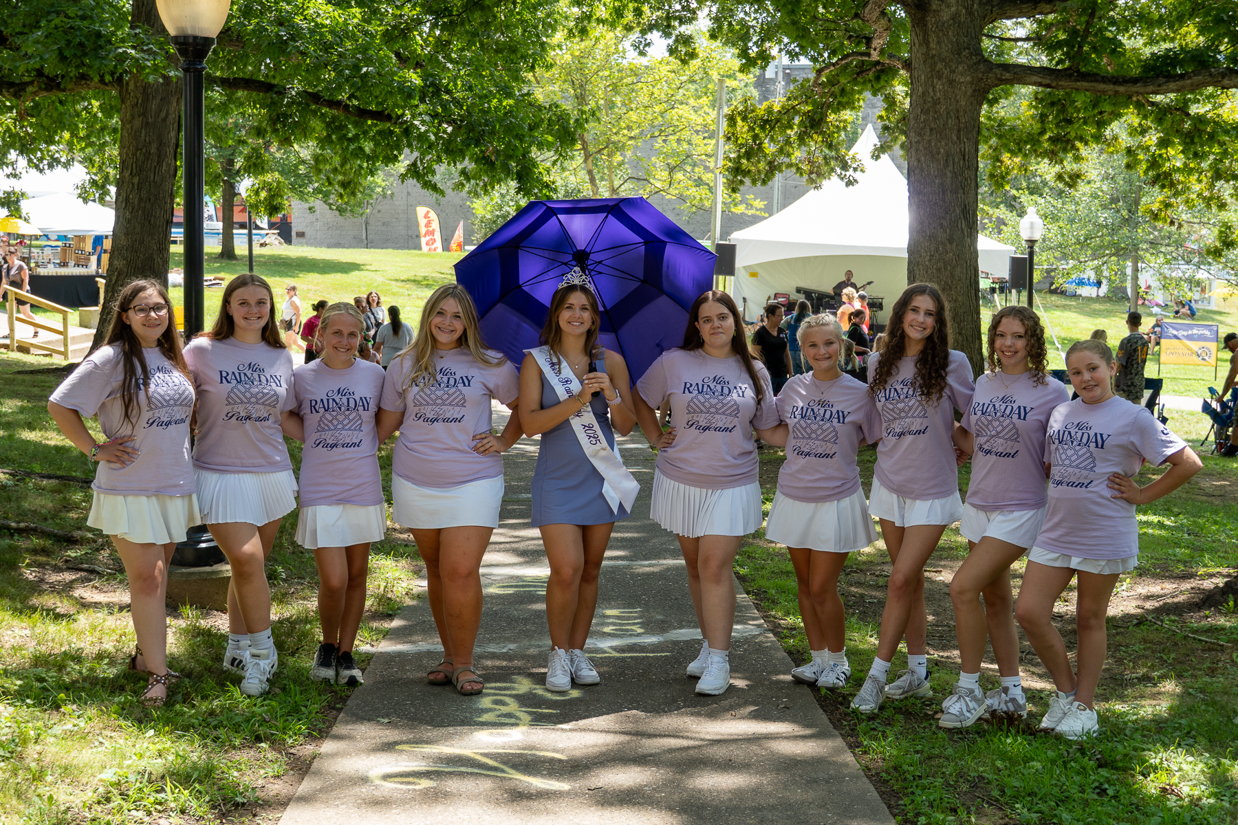 A group of girls wearing matching light purple “Miss Rain Day Pageant” shirts and white skirts stand together on a park path, posing around a crowned pageant winner holding a purple umbrella at an outdoor festival.