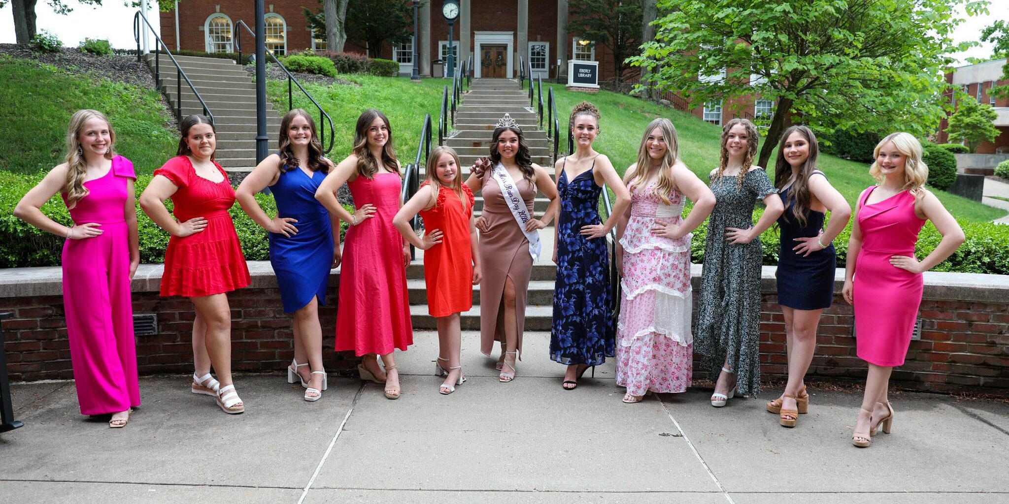 Group of young women in colorful formal dresses posing outdoors on a walkway in front of a brick building with columns and steps; one in the center wears a crown and sash.