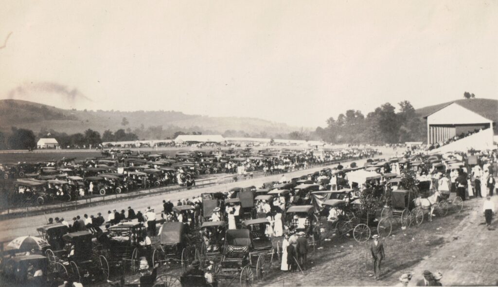 Historic black-and-white photo of a large outdoor gathering at a racetrack, with rows of early automobiles and horse-drawn carriages parked beside a grandstand filled with spectators and crowds lining the track.