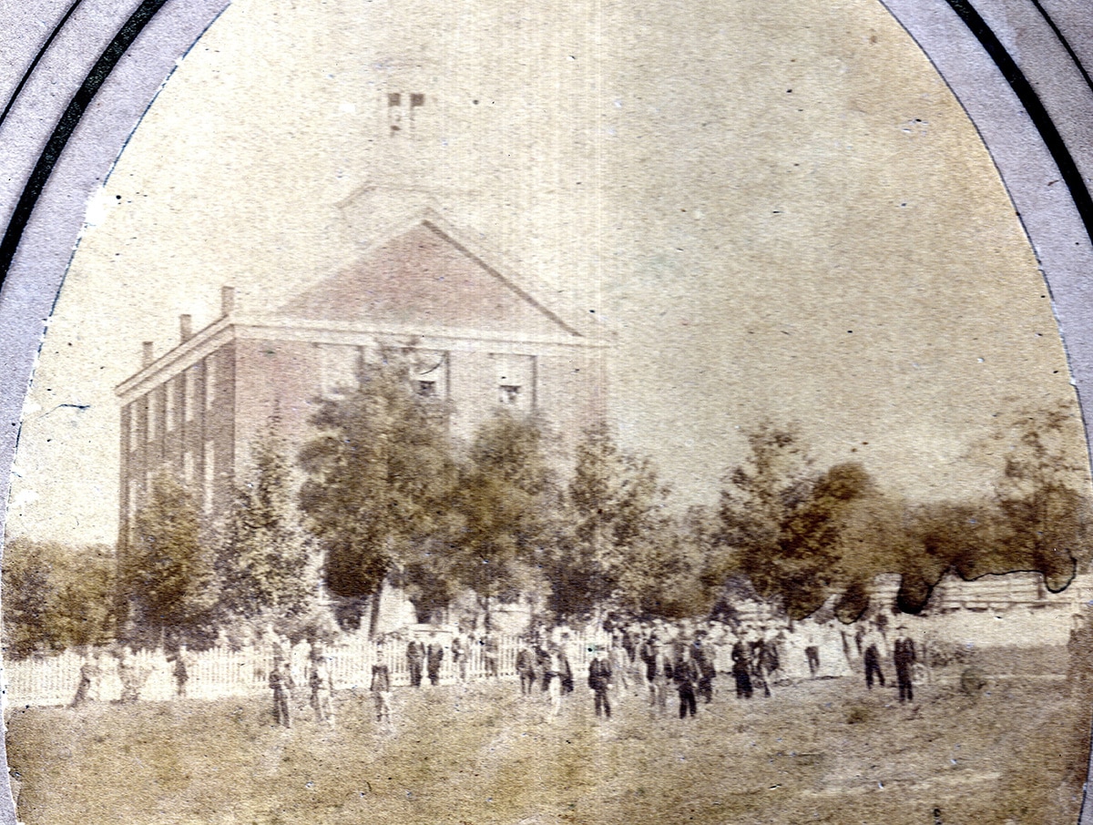 Faded historic photograph of a large columned building partially obscured by trees, with a crowd of people gathered in an open grassy area in front.