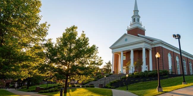 A scenic view of Waynesburg University's Robert Chapel with trees in the foreground and sunlight casting long shadows across the sidewalk. The chapel features a tall steeple, columns, and large windows, set against a clear blue sky.