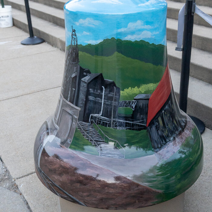 Painted bell sculpture featuring a historic coal mine, green hills, blue sky, and mining structures on display outdoors.