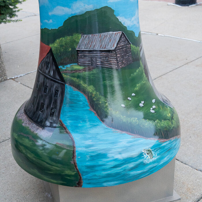 Painted bell sculpture featuring a rustic log cabin beside a winding blue stream, with green hills, wildflowers, and a clear blue sky, displayed as part of the Bells Across PA public art exhibit.