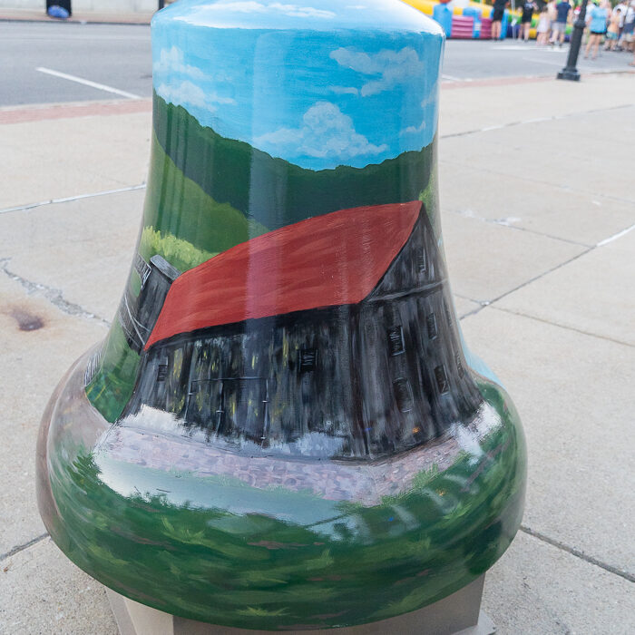 Painted bell sculpture featuring a historic wooden barn with a red roof set against rolling green hills and a blue sky, displayed as part of the Bells Across PA public art exhibit.