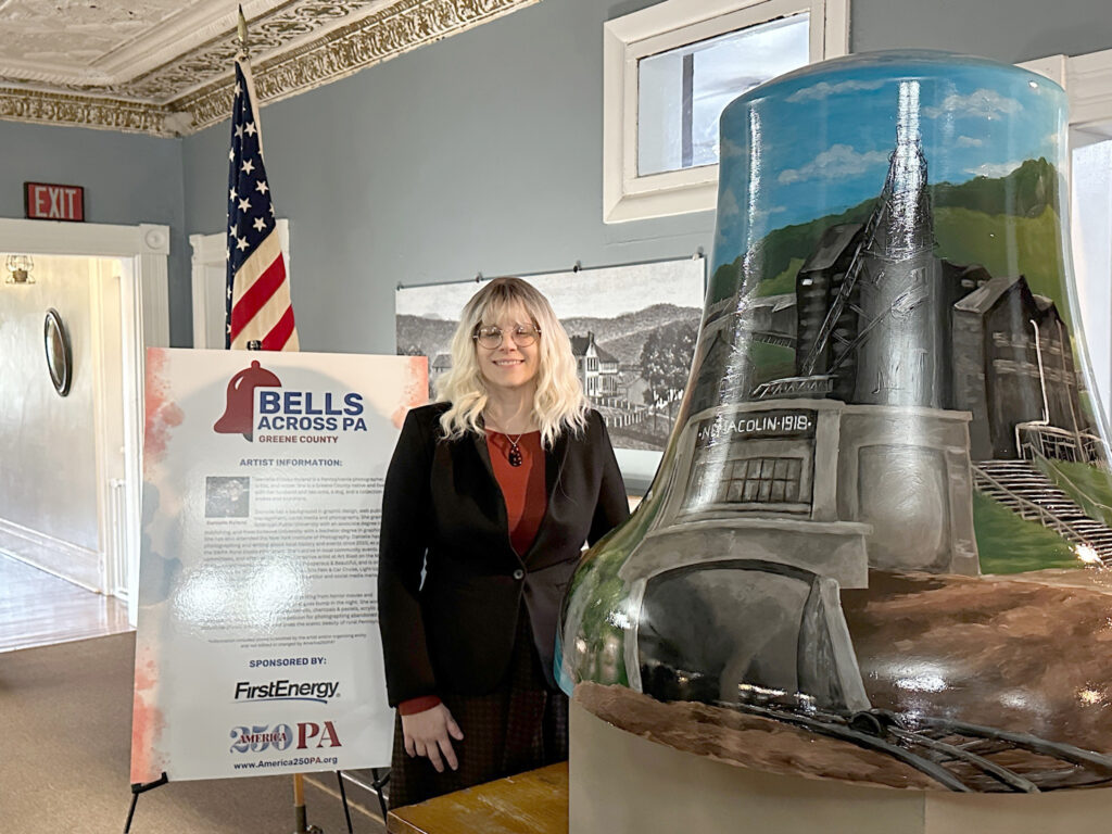 Woman standing beside a large painted bell sculpture featuring an industrial mining scene, displayed indoors at a “Bells Across PA – Greene County” exhibit. An American flag and informational sign are visible in the background of the historic-style room.