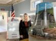 Woman standing beside a large painted bell sculpture featuring an industrial mining scene, displayed indoors at a “Bells Across PA – Greene County” exhibit. An American flag and informational sign are visible in the background of the historic-style room.