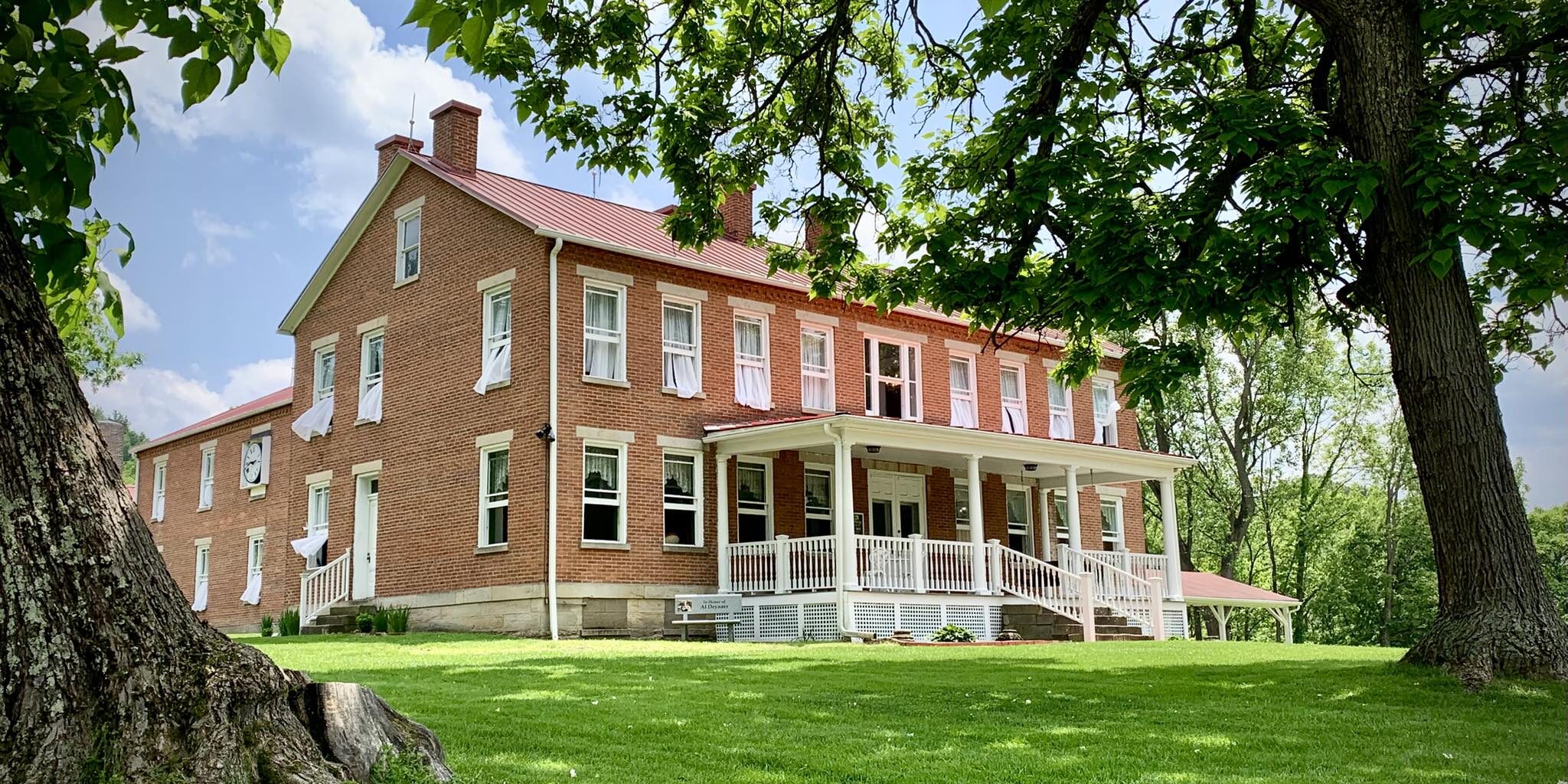 Historic two-story red brick building with a white wraparound porch, surrounded by large shade trees and a manicured green lawn on a sunny day.