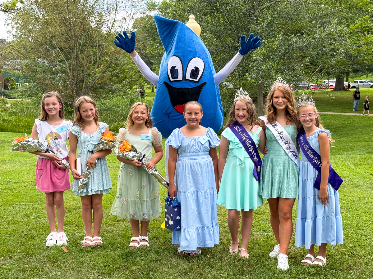 Contestants and pageant winners pose with a large blue raindrop mascot during the Miss Rain Day Pageant in a park.