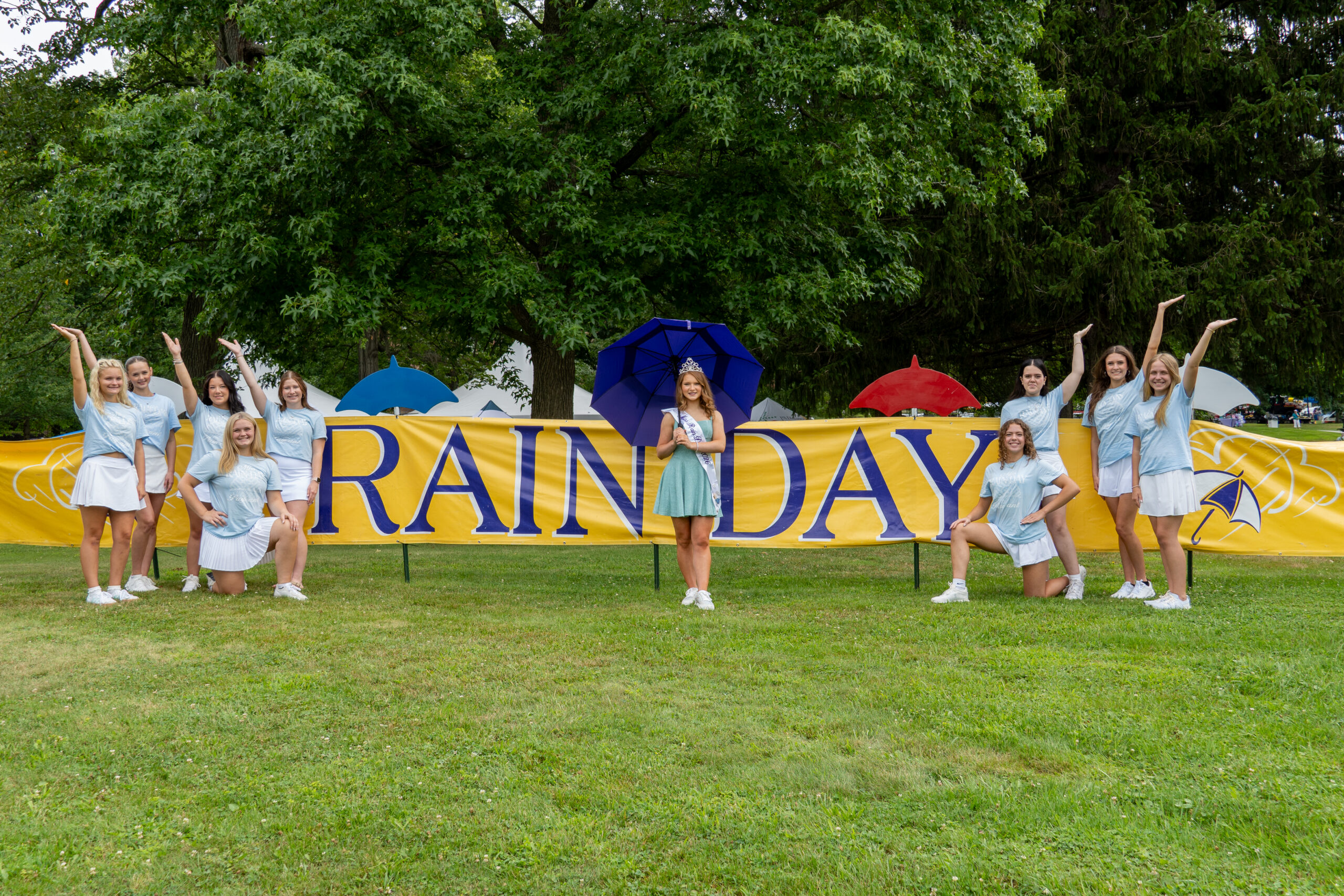The reigning Miss Rain Day and pageant contestants pose outdoors in front of a large "Rain Day" banner, with colorful umbrellas and trees in the background.