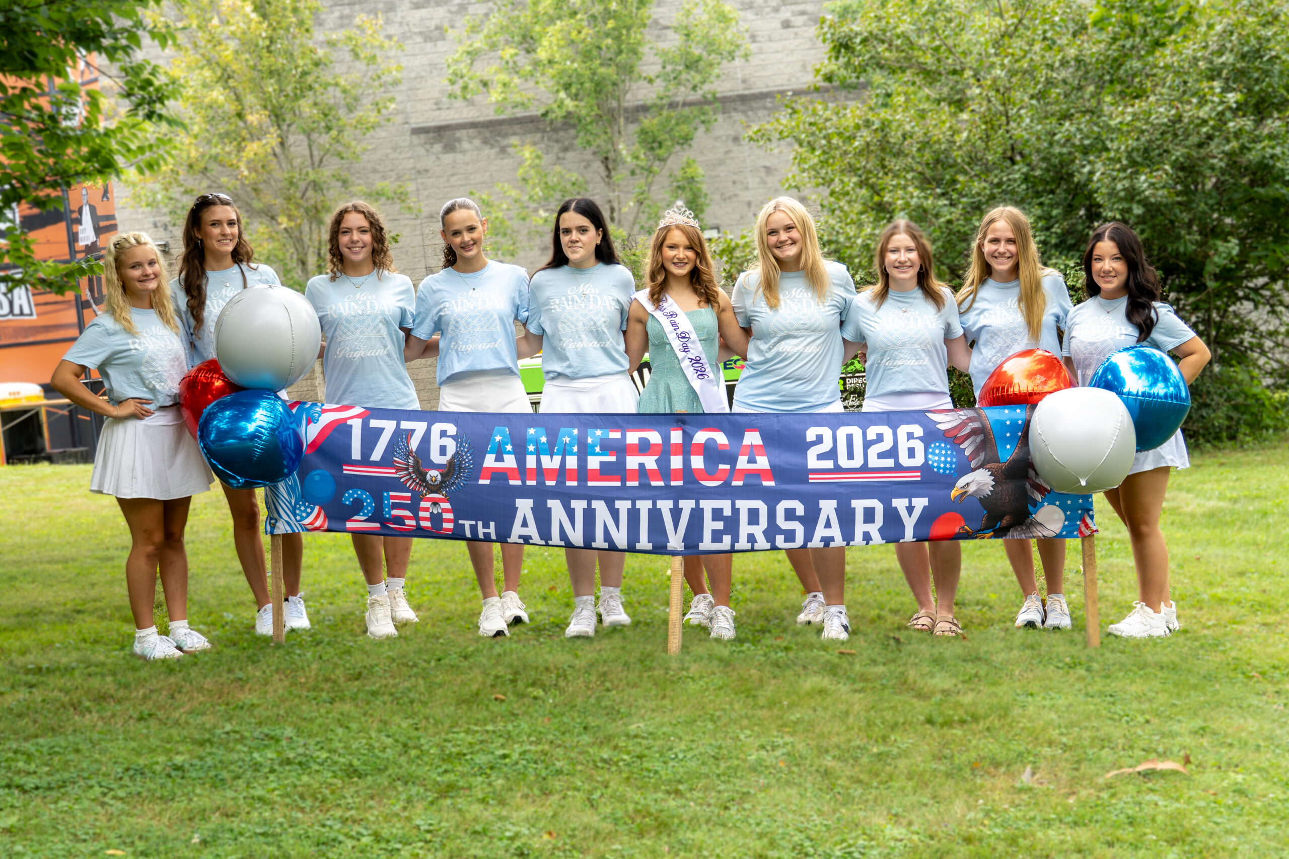 A group of pageant contestants and the reigning Miss Rain Day pose outdoors behind a patriotic "America 2026 – 250th Anniversary" banner with red, white, and blue balloons.