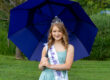 Young pageant participant wearing a tiara and "Miss Rain Day 2026" sash, holding a blue umbrella outdoors.