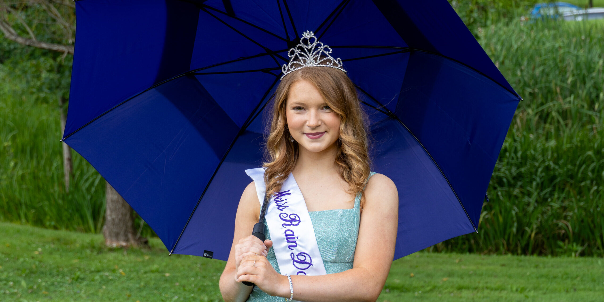 20260729 Miss Rain Day and Contestants-33 Young pageant participant wearing a tiara and "Miss Rain Day 2026" sash, holding a blue umbrella outdoors.