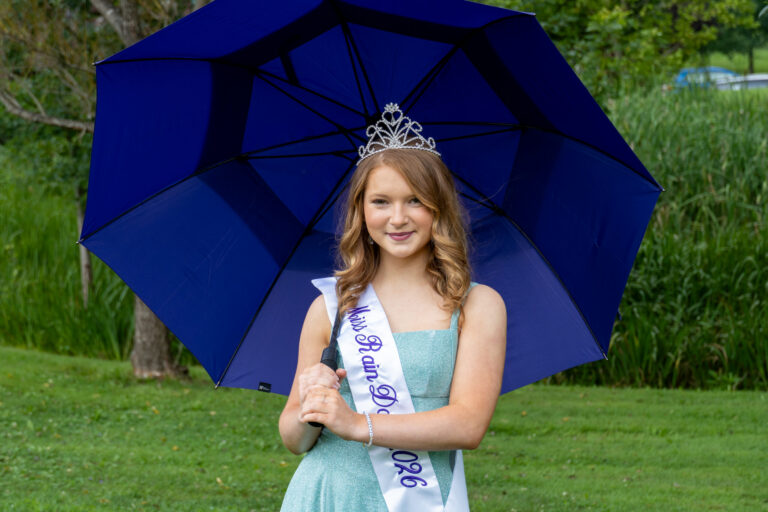 20260729 Miss Rain Day and Contestants-33 Young pageant participant wearing a tiara and "Miss Rain Day 2026" sash, holding a blue umbrella outdoors.
