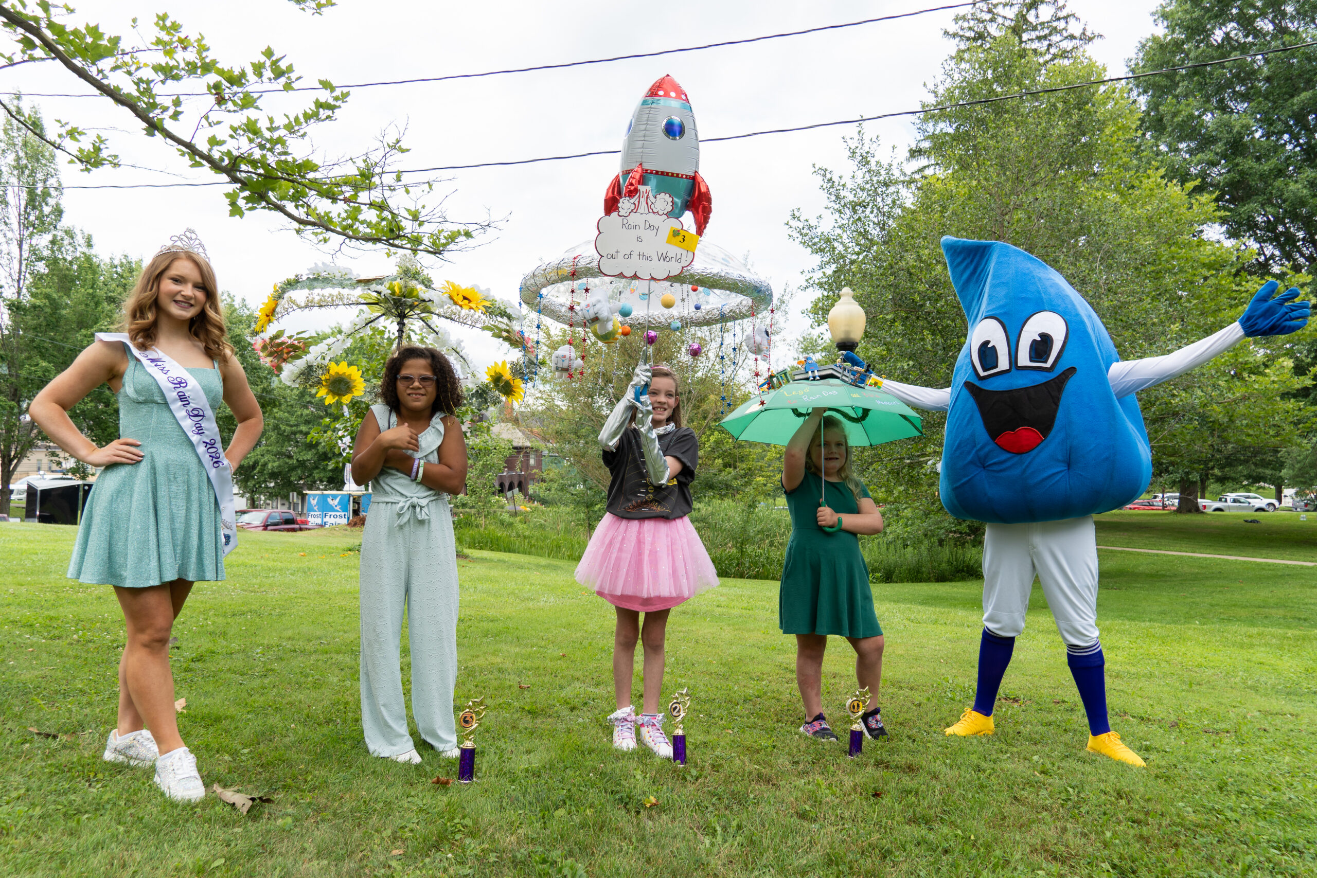 Miss Rain Day stands with children displaying decorated umbrellas alongside the Rain Day mascot during the Rain Day Festival umbrella contest.