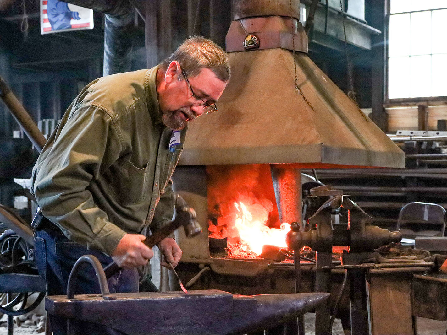 A blacksmith in a workshop hammers a glowing piece of metal on an anvil. Flames blaze in the forge behind him, and various tools and equipment are scattered around the workspace.