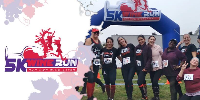 Group of women posing under a ‘5K Wine Run – Run Now, Wine Later’ finish arch, wearing race bibs and athletic outfits, smiling at an outdoor event.